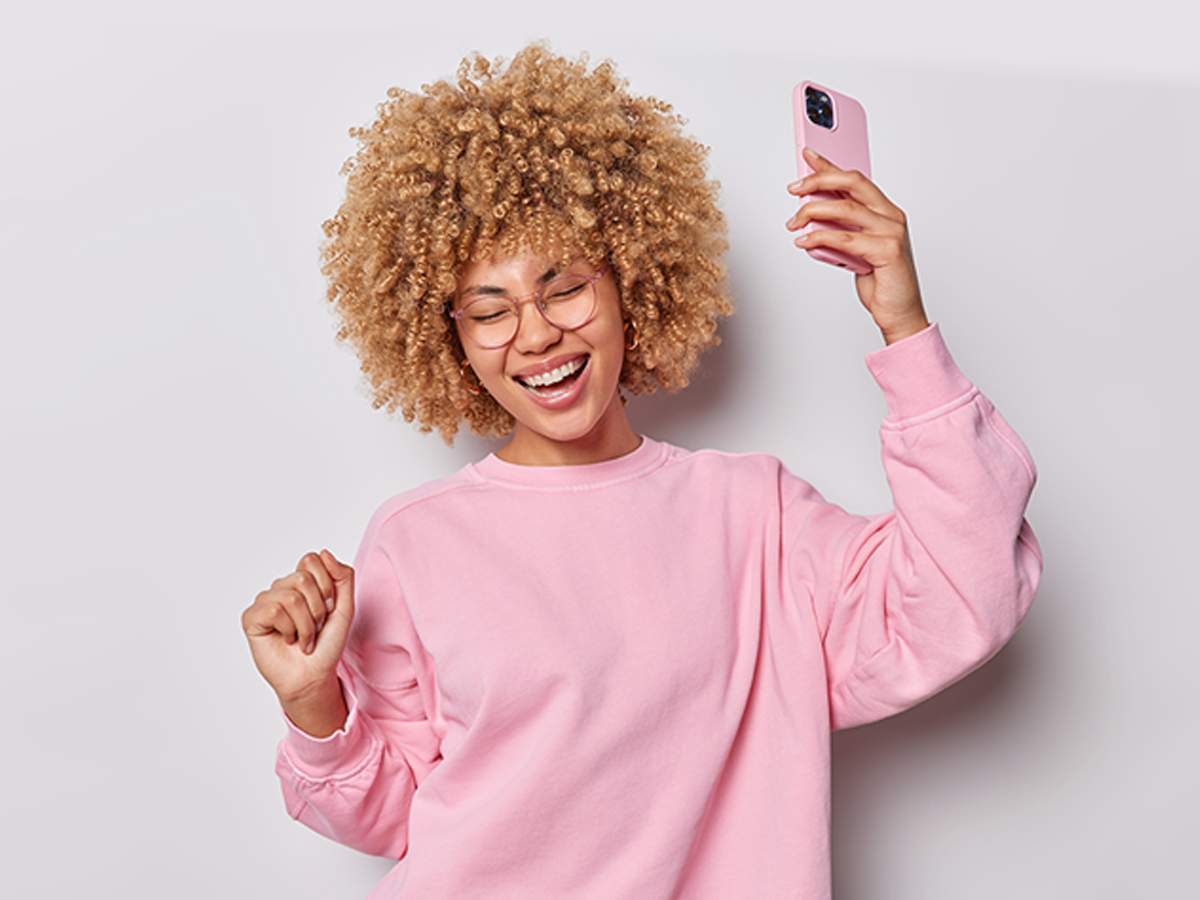 Woman with coily hair holding cell phone up while smiling